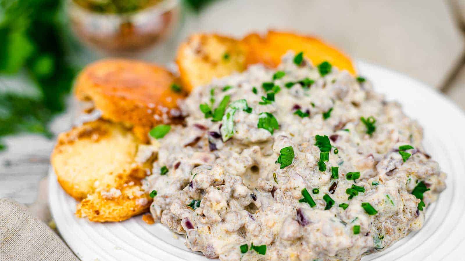 A plate of creamy sausage gravy garnished with chopped green herbs is served alongside golden-brown toasted bread slices. The dish appears rich and hearty, with visible bits of sausage and herbs mixed into the gravy.