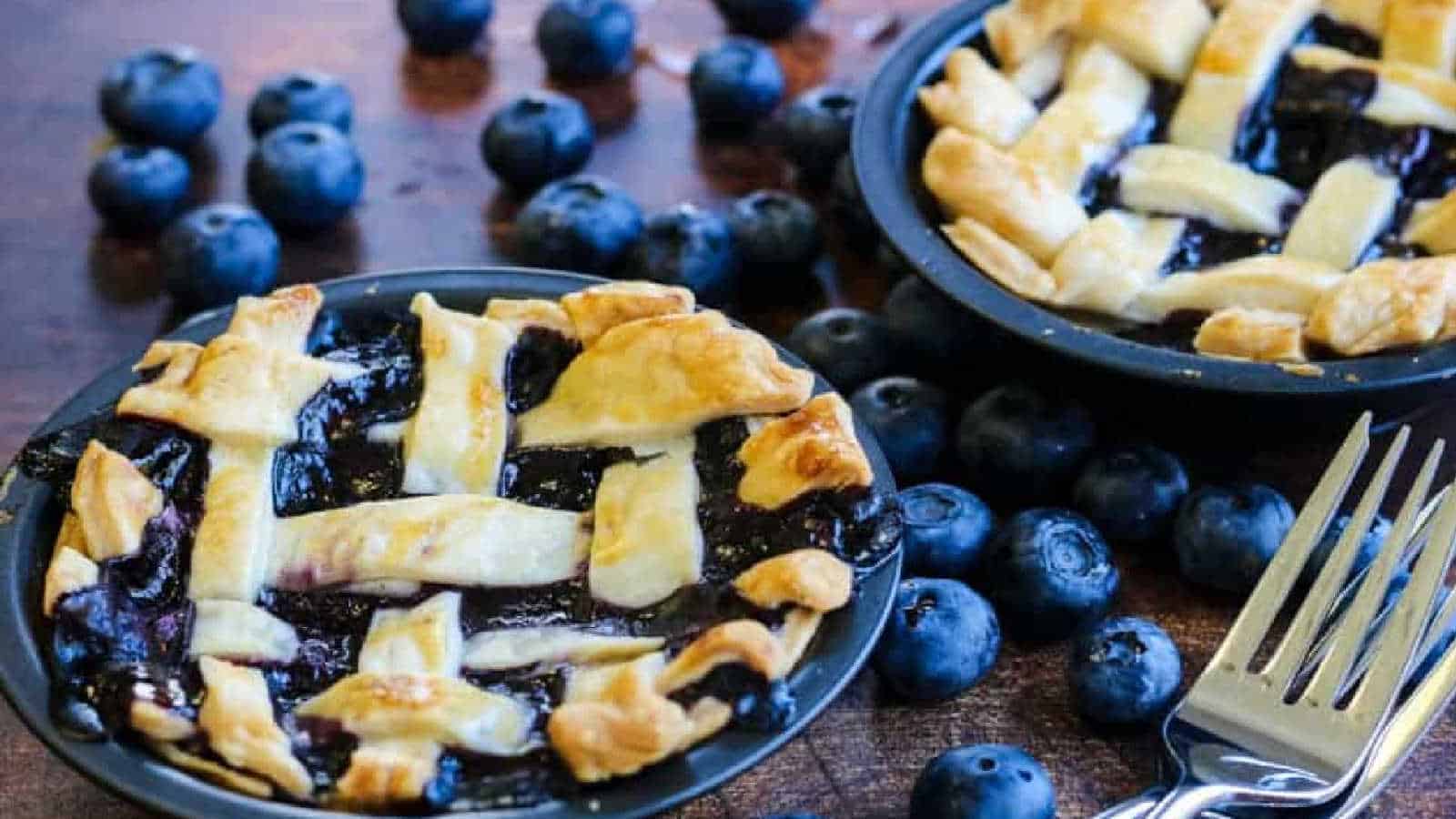 Two small blueberry pies with golden lattice crusts sit on a wooden surface, surrounded by fresh blueberries. A fork rests nearby, ready for use. The vibrant blueberries contrast with the rustic brown background, creating a visually appealing scene.