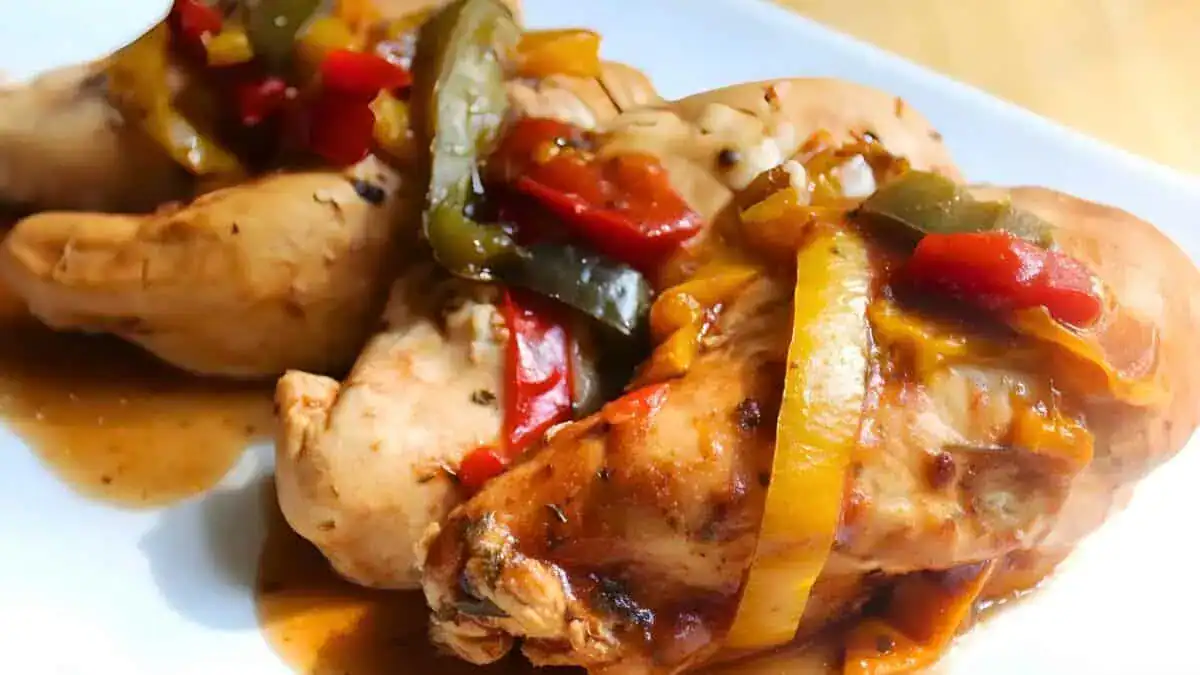 A close-up of a plate with three seasoned and cooked chicken breasts, topped with sautéed green and red bell peppers and onions, all drizzled with a savory sauce. The dish appears vibrant and flavorful.