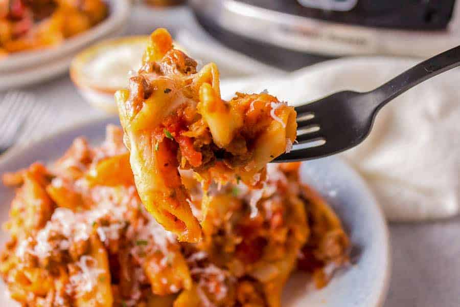 A black fork holds a bite of pasta topped with a hearty meat sauce, with the rest of the pasta dish visible on a white plate in the background. The pasta has a sprinkling of grated cheese and herbs, and the scene features a slow cooker in the distant background.