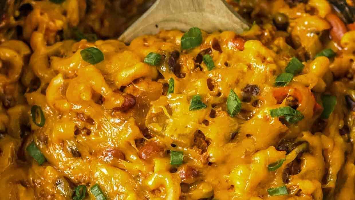 A close-up view of a cheesy, baked pasta dish with various vegetables mixed in. The golden melted cheese blankets the pasta, and chopped green onions garnish the top, adding a pop of color and freshness. A wooden spoon is partially visible in the background.