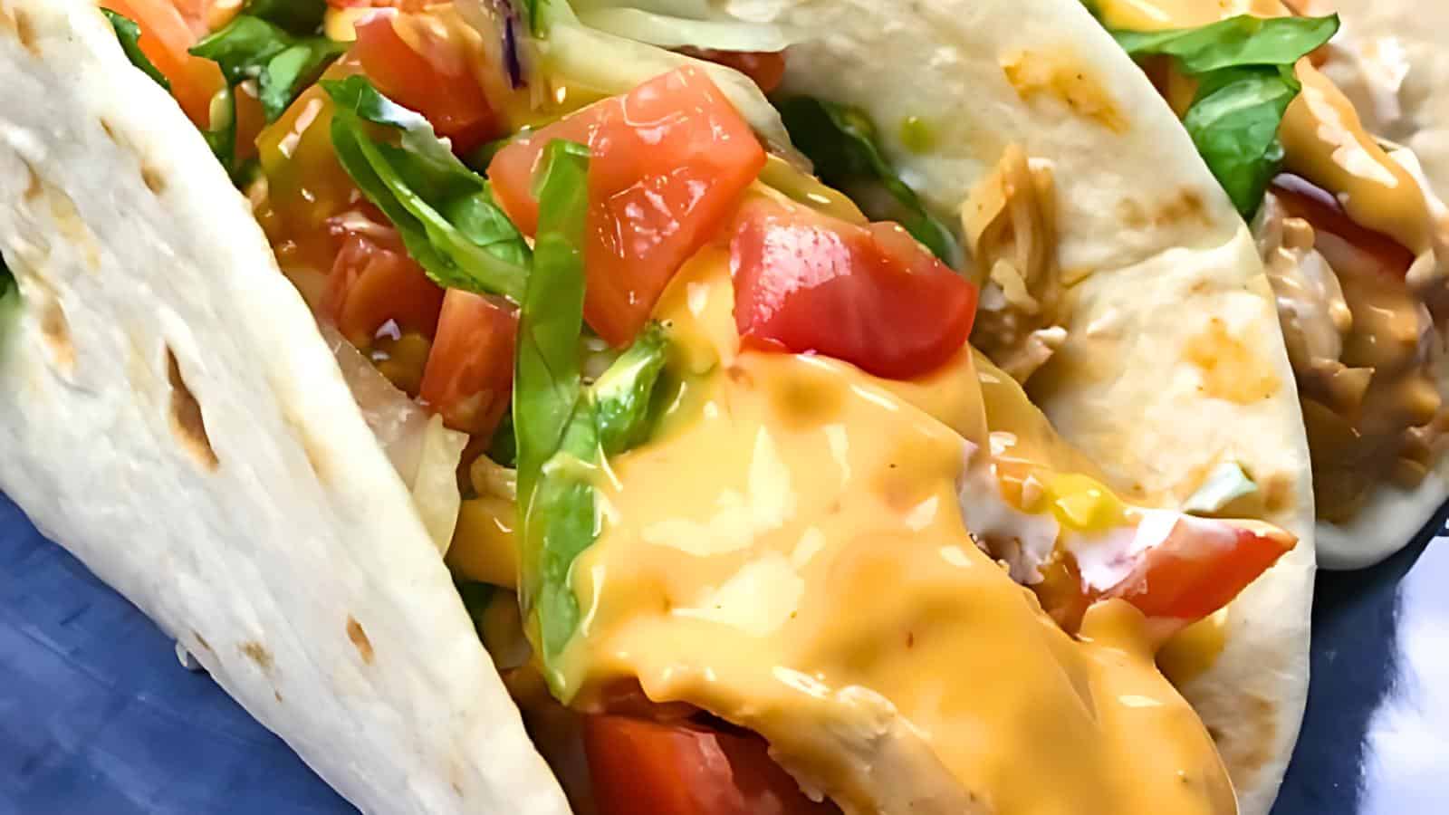 Close-up of two soft tacos filled with diced tomatoes, shredded lettuce, and melted cheese sauce on a blue plate.