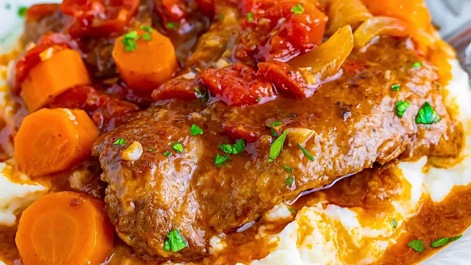 A close-up image of a savory beef stew on a bed of mashed potatoes. The stew includes chunks of carrots, diced tomatoes, and onions in a rich, reddish-brown sauce, garnished with chopped parsley. The creamy mashed potatoes provide a comforting base for the hearty dish.