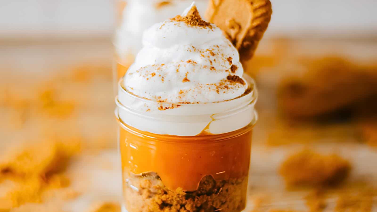 A small mason jar filled with a layered dessert including a crumbled base, a smooth caramel-colored middle layer, and topped with fluffy whipped cream. A cookie is partially inserted into the whipped cream, and a sprinkle of spices is visible on top.