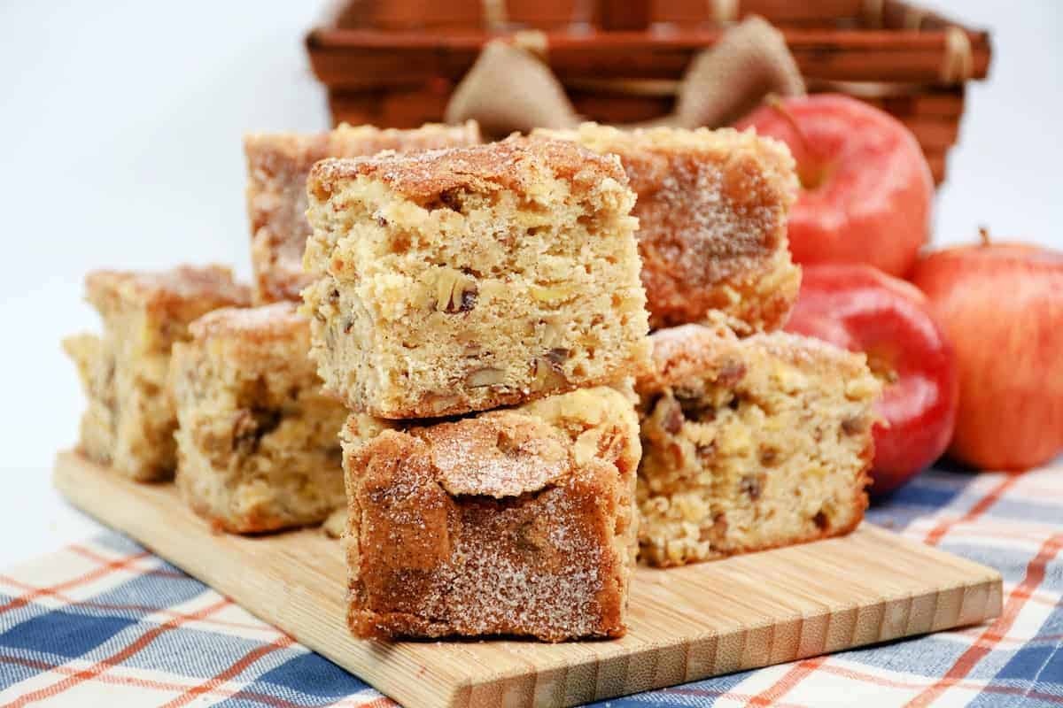 A wooden cutting board holds several thick slices of apple cake topped with a sugar crust. The background includes a checkered blue and red cloth and some whole red apples, with a woven basket behind them. The scene is brightly lit, emphasizing the dessert’s texture.