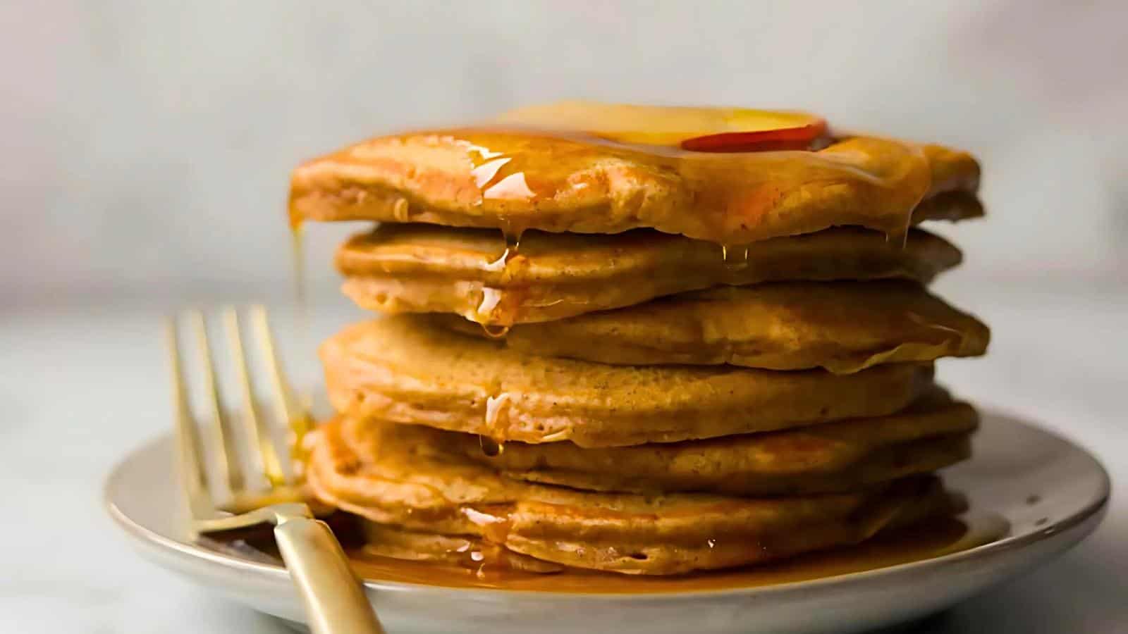 A stack of golden-brown pancakes drenched in syrup, topped with a pat of melting butter and a thin slice of fruit, sits on a white plate with a gold-colored fork resting beside it. The background is softly blurred, emphasizing the main focus on the pancakes.