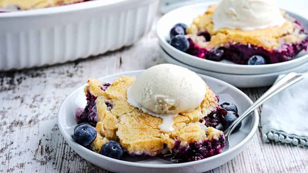 Close-up of two plates of blueberry cobbler topped with a scoop of vanilla ice cream. The cobbler has a golden-brown crust and fresh blueberries are visible. A white baking dish and a napkin can be seen in the background on a rustic wooden table.