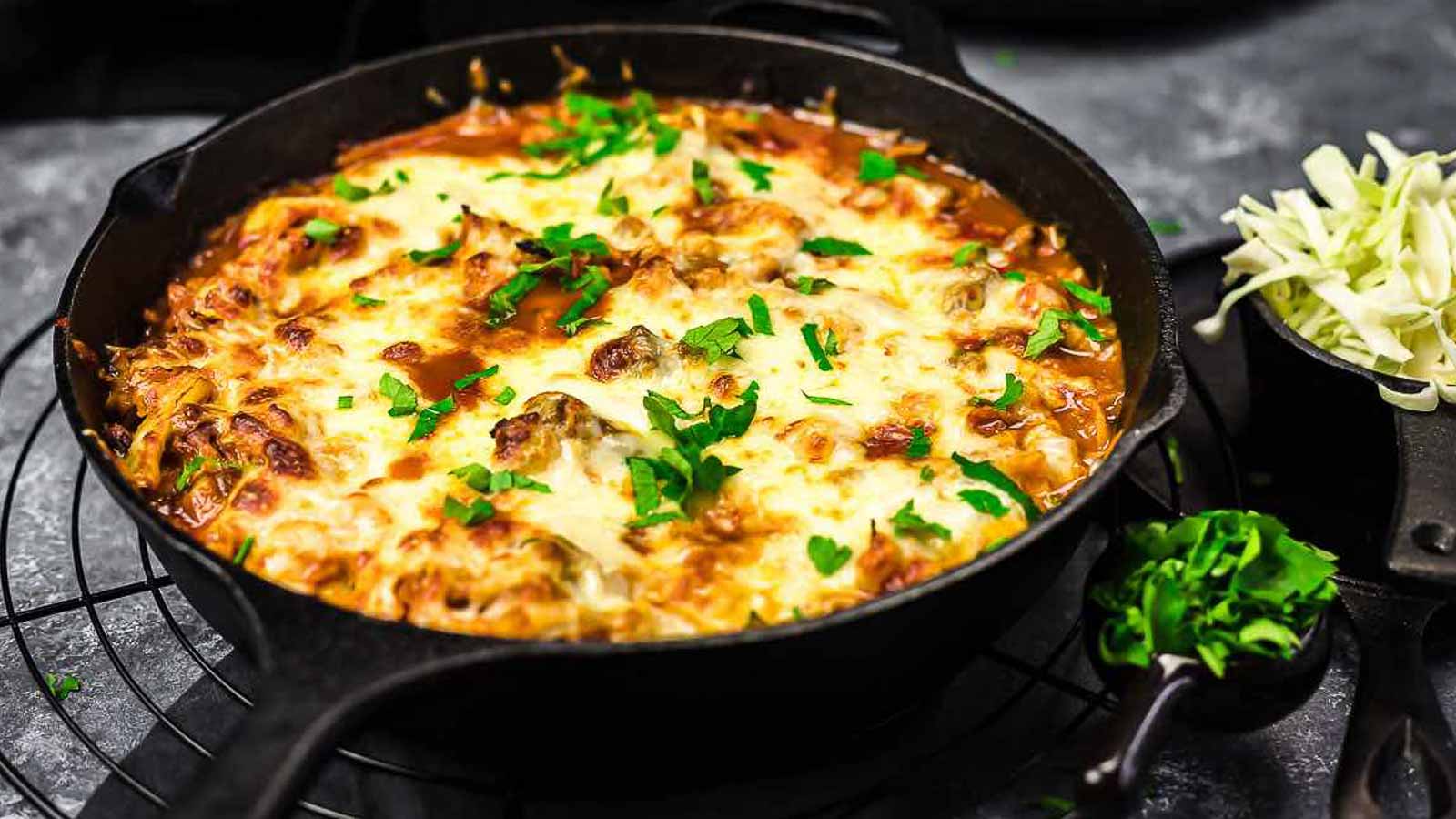 A cast iron skillet filled with a baked dish topped with melted cheese and garnished with chopped parsley. The dish appears rich and hearty, with a small bowl of shredded cheese and another of chopped herbs nearby. The skillet is placed on a wire rack.