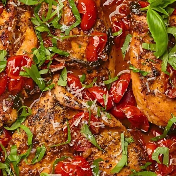 A close-up of a skillet containing cooked chicken breasts, cherry tomatoes, fresh basil, and grated Parmesan cheese.
