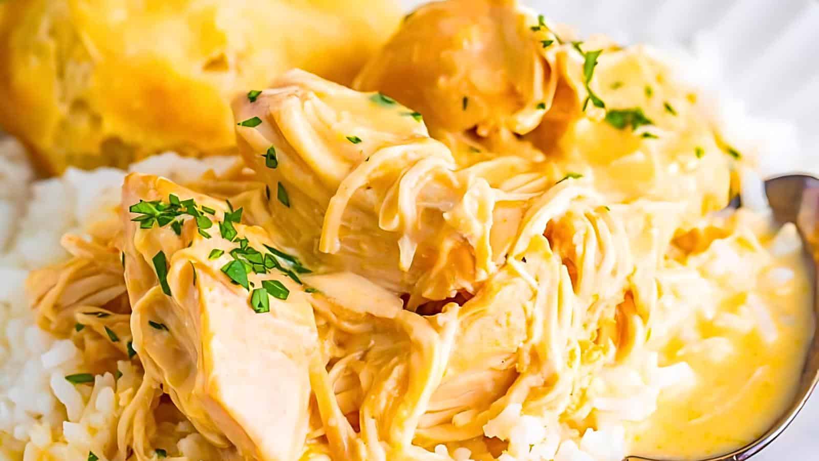 A close-up of shredded chicken in a creamy yellow sauce served over white rice, garnished with chopped parsley, and accompanied by a piece of bread.