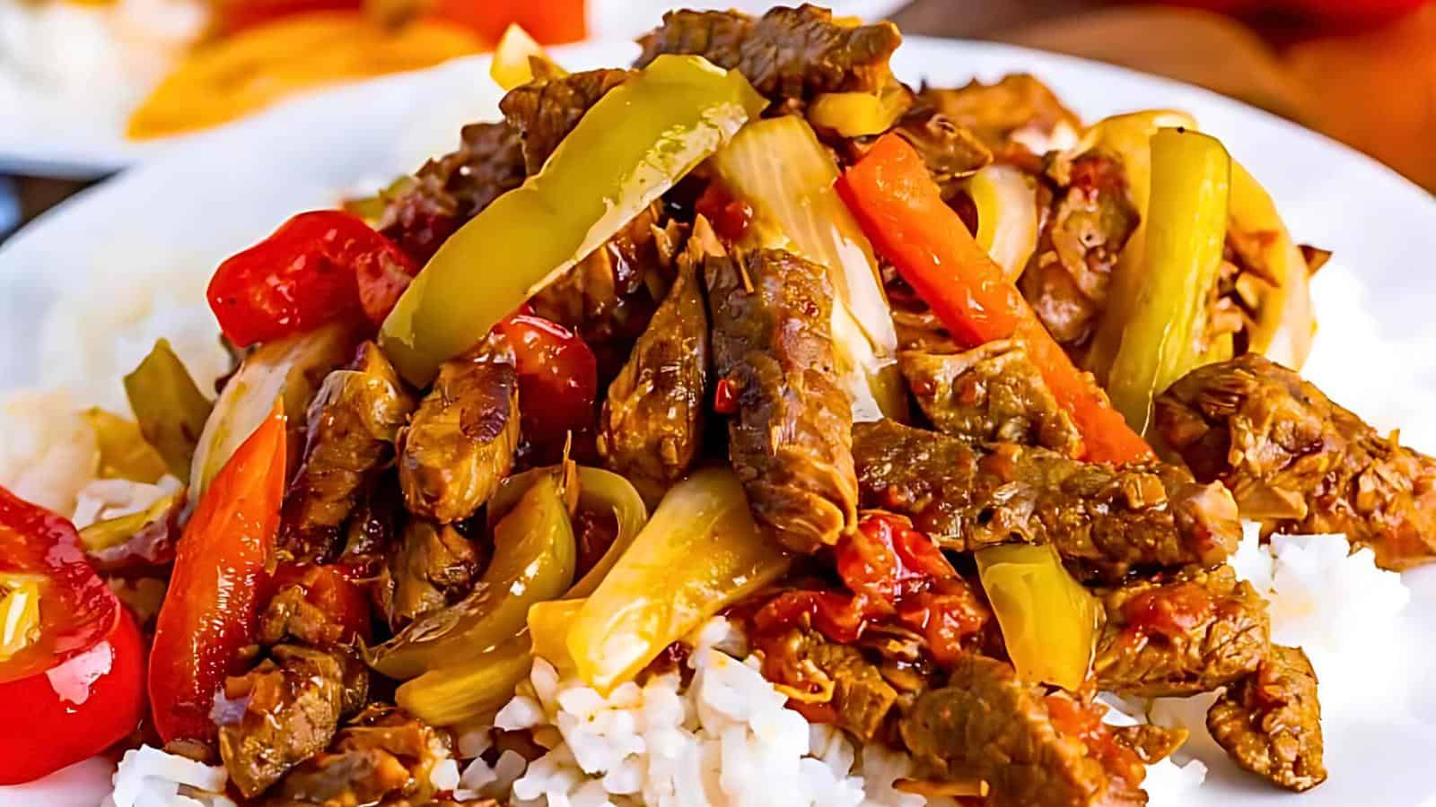 A plate of beef stir-fry is served with a bed of white rice. The dish features tender beef strips, vibrant bell peppers in red and green, and sliced onions, all coated in a savory sauce. The colorful vegetables and rich beef are appetizingly arranged atop the rice.