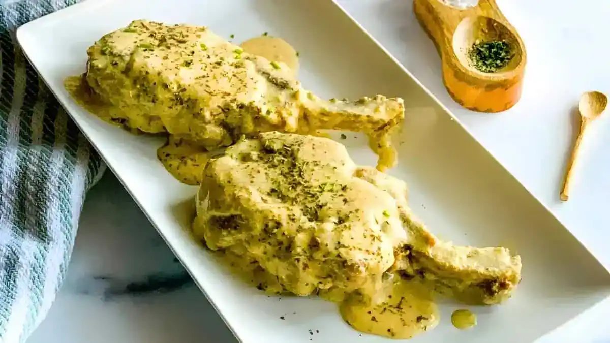 A rectangular white plate features two smothered pork chops, coated in a creamy herb sauce and garnished with dried herbs. A spoonful of dried herbs in a wooden dish with a tiny wooden spoon is placed in the background, alongside a folded striped cloth.