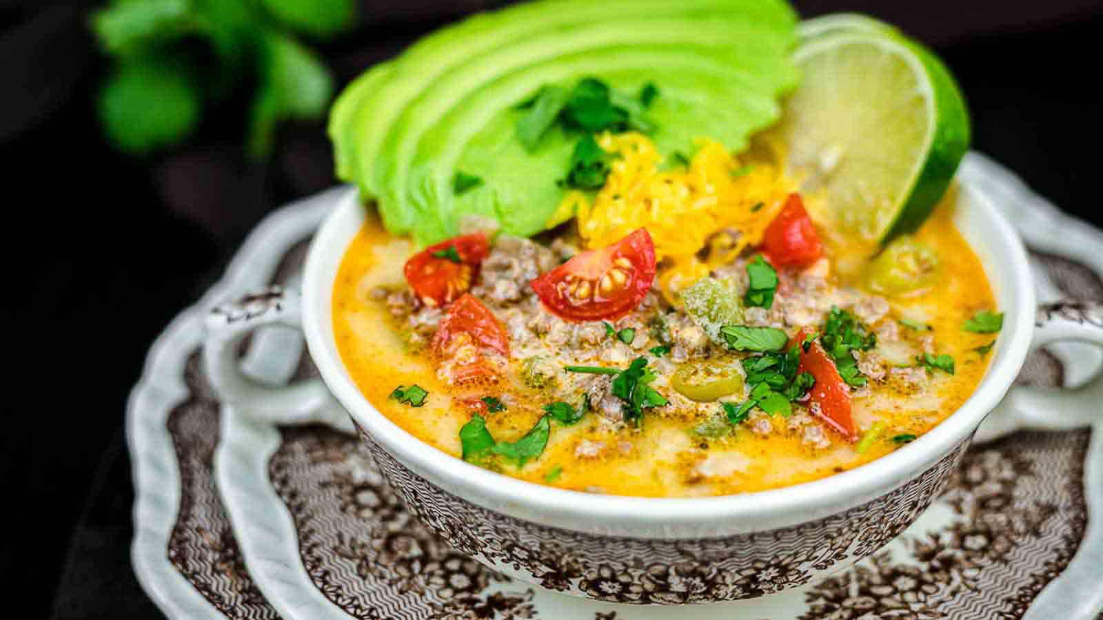 A bowl of creamy soup garnished with slices of avocado, lime, shredded cheese, chopped tomatoes, and cilantro. The bowl is placed on a decorative plate, with a dark background and a hint of green foliage on the side.