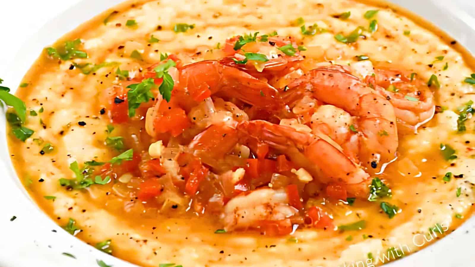 A close-up of a plate of shrimp and grits. The creamy grits are topped with cooked shrimp, diced tomatoes, a savory sauce, and garnished with fresh herbs and ground black pepper.