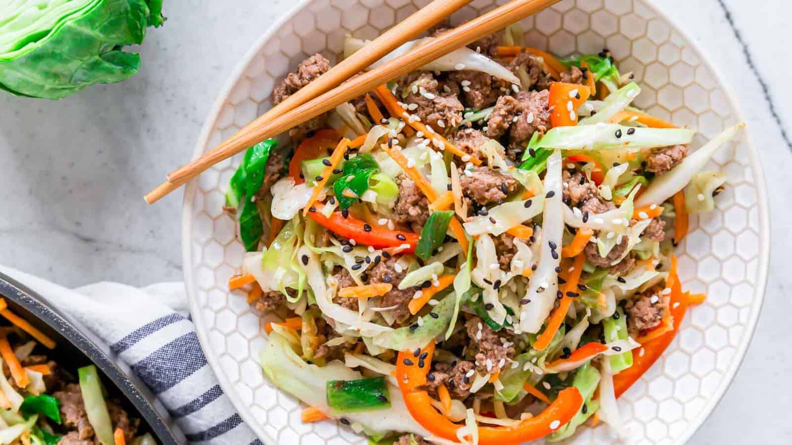 A bowl of vibrant stir-fried vegetables, including shredded cabbage, carrot, and bell pepper, mixed with ground meat and garnished with sesame seeds. A pair of wooden chopsticks rests on the edge of the bowl.