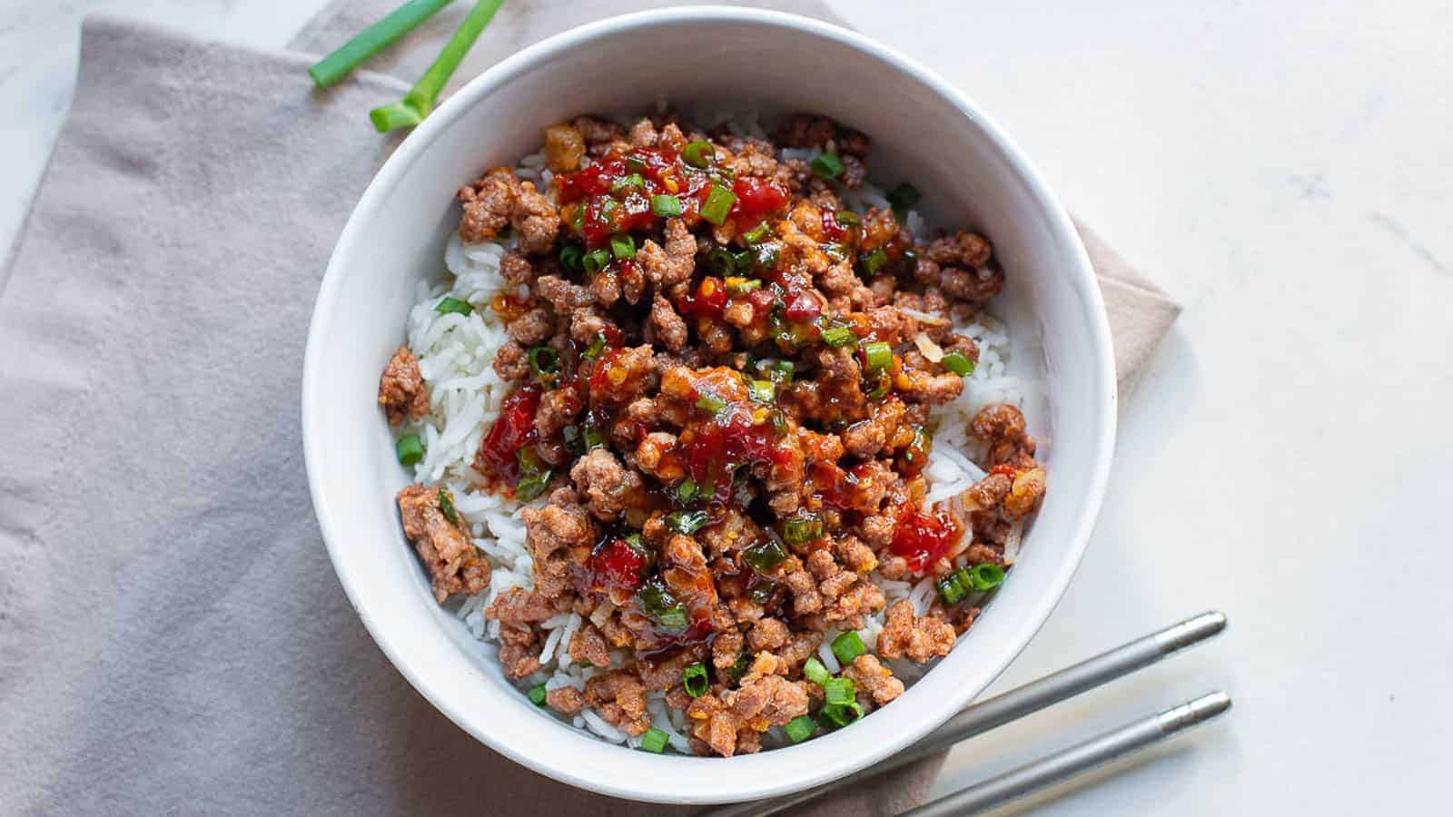 A white bowl filled with white rice topped with minced meat, chopped green onions, and a sauce. Two metal chopsticks are placed on the side of the bowl, resting on a beige napkin. Green onion stalks are visible in the background.