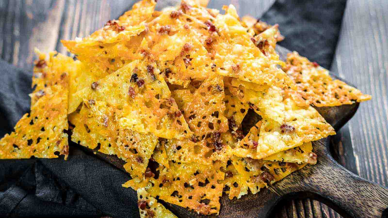 A pile of crispy, golden-brown cheese crisps with sprinkled bacon bits are arranged on a wooden board placed over a dark cloth. The background features a rustic wooden table. The crisps have a textured, lattice-like appearance.