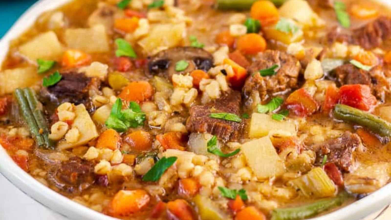 A bowl filled with hearty beef and barley soup, featuring chunks of tender beef, diced potatoes, sliced carrots, green beans, mushrooms, and barley. The soup is topped with fresh parsley and appears rich and savory with a variety of vibrant ingredients.