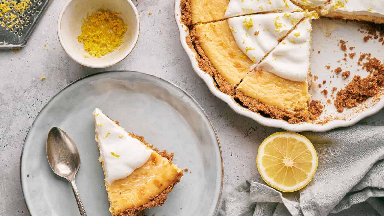 A partially sliced lemon pie with a graham cracker crust and whipped topping in a pie dish, a lemon half, and zest on a gray cloth. A plate with a slice of the pie and a silver spoon is set beside it, along with a small bowl of grated lemon zest and a grater.