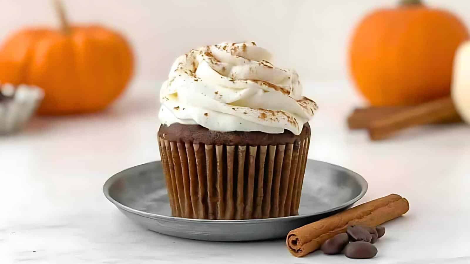 A chocolate cupcake topped with white frosting and a sprinkle of cinnamon sits on a small metal plate. In the background are two pumpkins and cinnamon sticks, enhancing the autumn theme. A couple of chocolate chips and a cinnamon stick are placed beside the plate.