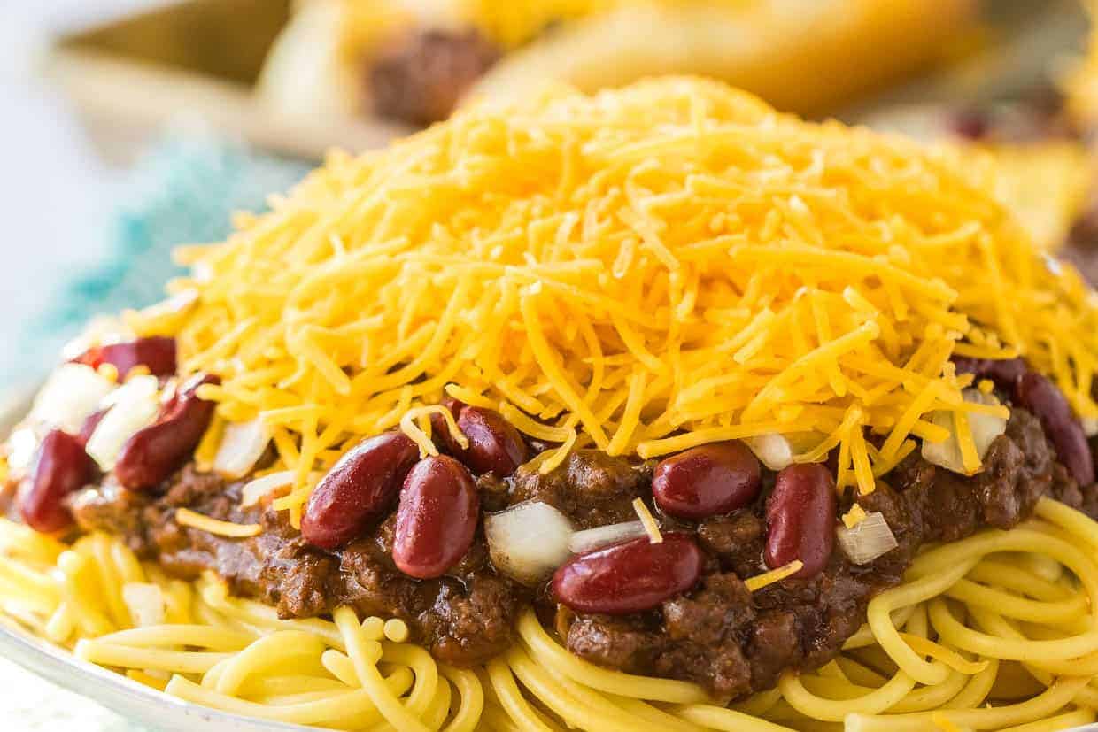 A plate of Cincinnati-style chili spaghetti is topped with kidney beans, finely chopped onions, and a generous mound of shredded cheddar cheese. The chili meat sauce is spread evenly over the spaghetti, offering a hearty and colorful presentation.