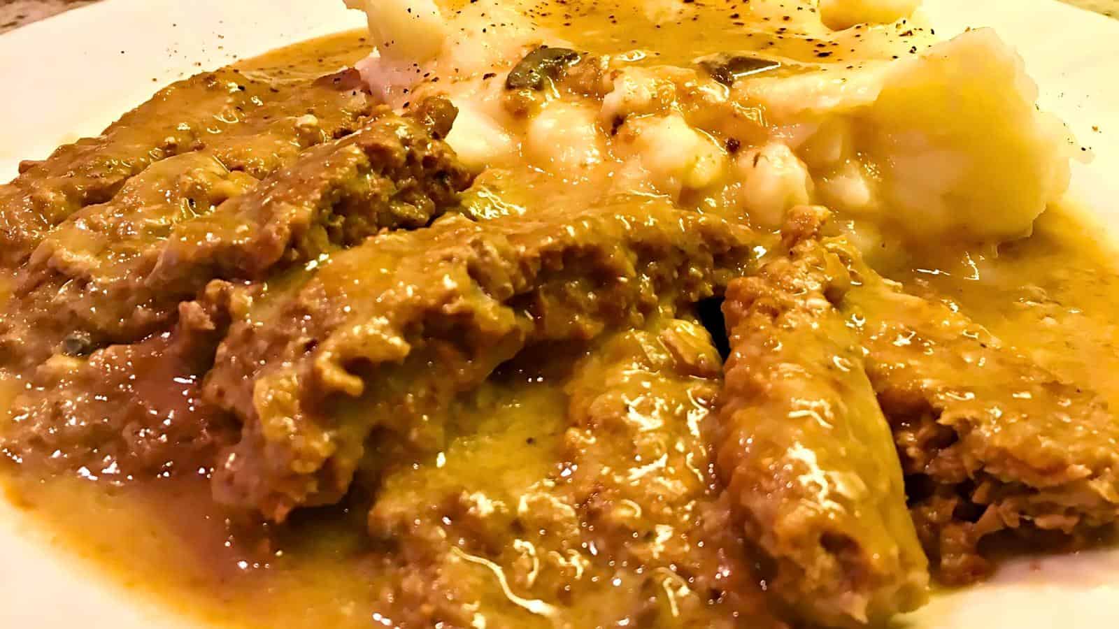 A close-up of a plate of sliced meatloaf covered in a thick, brown gravy, with a serving of mashed potatoes topped with a sprinkle of black pepper beside it. The dish appears rich and hearty, ready to be enjoyed.