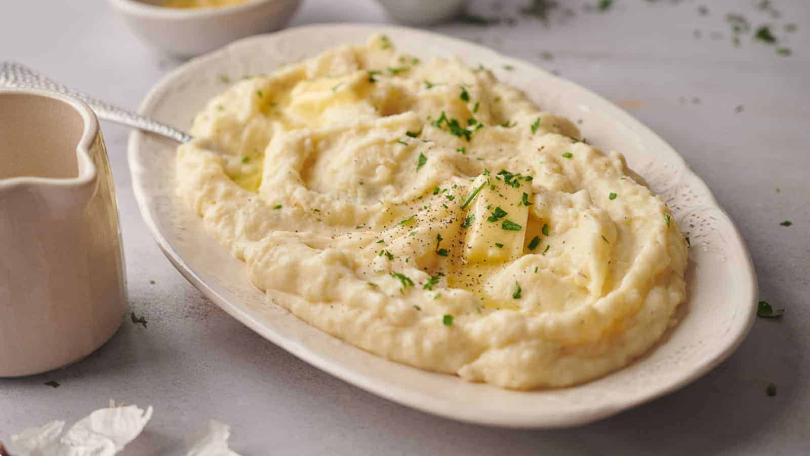 A white oval dish filled with creamy mashed potatoes, topped with small pieces of melting butter and garnished with chopped fresh herbs. A small pitcher of gravy is visible on the left side, and blurred background elements suggest a kitchen setting.