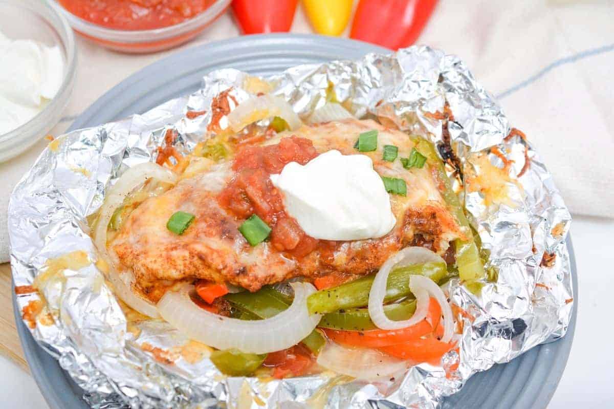 A foil packet containing a baked dish with melted cheese, onions, bell peppers, and chicken, topped with salsa, sour cream, and chopped green onions. The foil rests on a gray plate, with bowls of salsa and sour cream and colorful peppers in the background.
