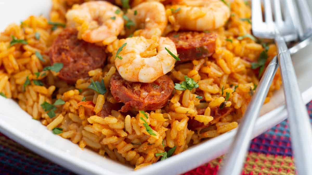A plate of jambalaya featuring seasoned rice, shrimp, and slices of sausage, garnished with chopped green herbs. A fork and a spoon rest on the side of the plate.