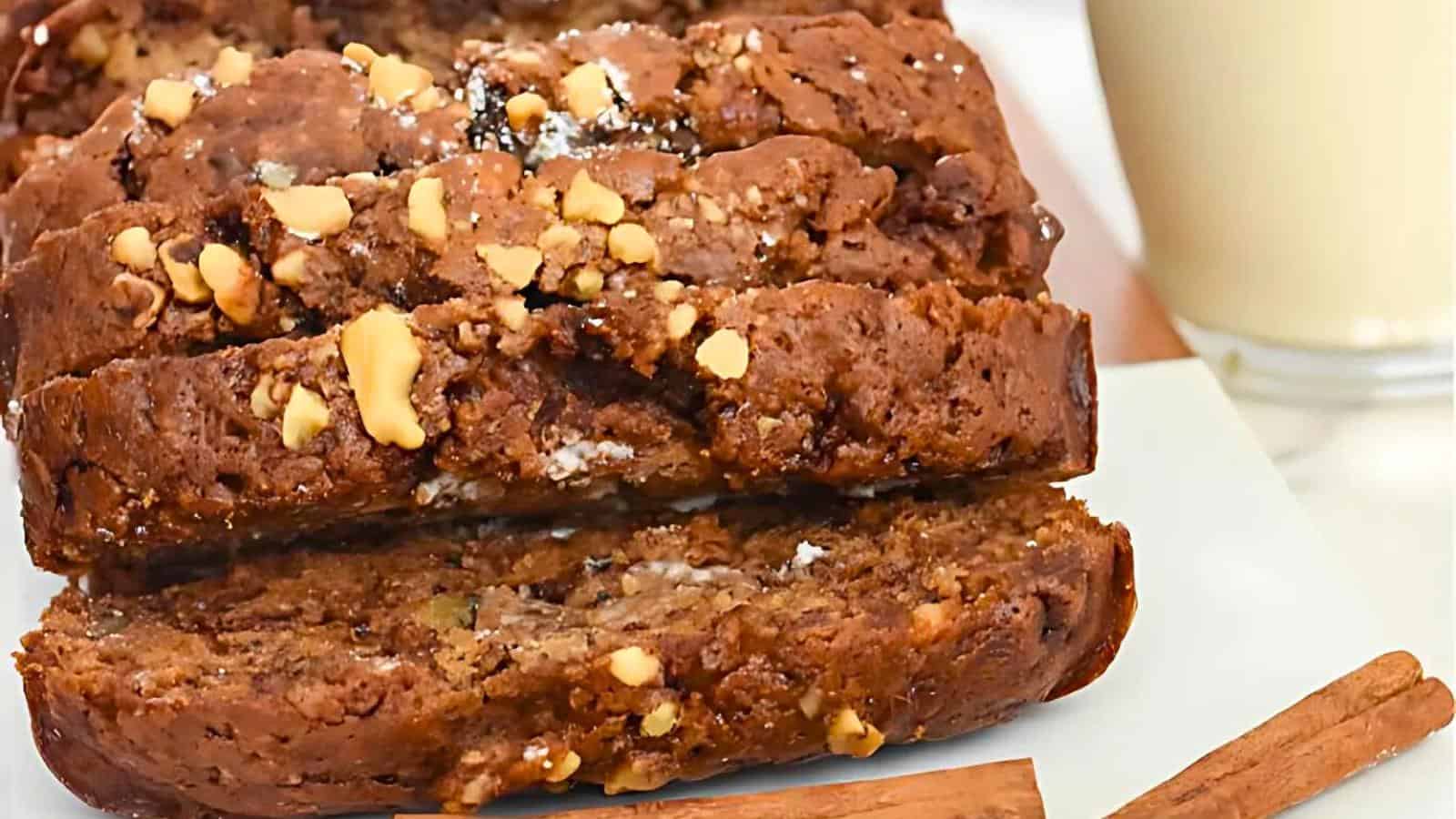 A close-up of sliced banana bread on a white plate. The bread is sprinkled with crushed walnuts and appears moist with visible chunks of banana. In the background, a glass of milk and a couple of cinnamon sticks are slightly out of focus.