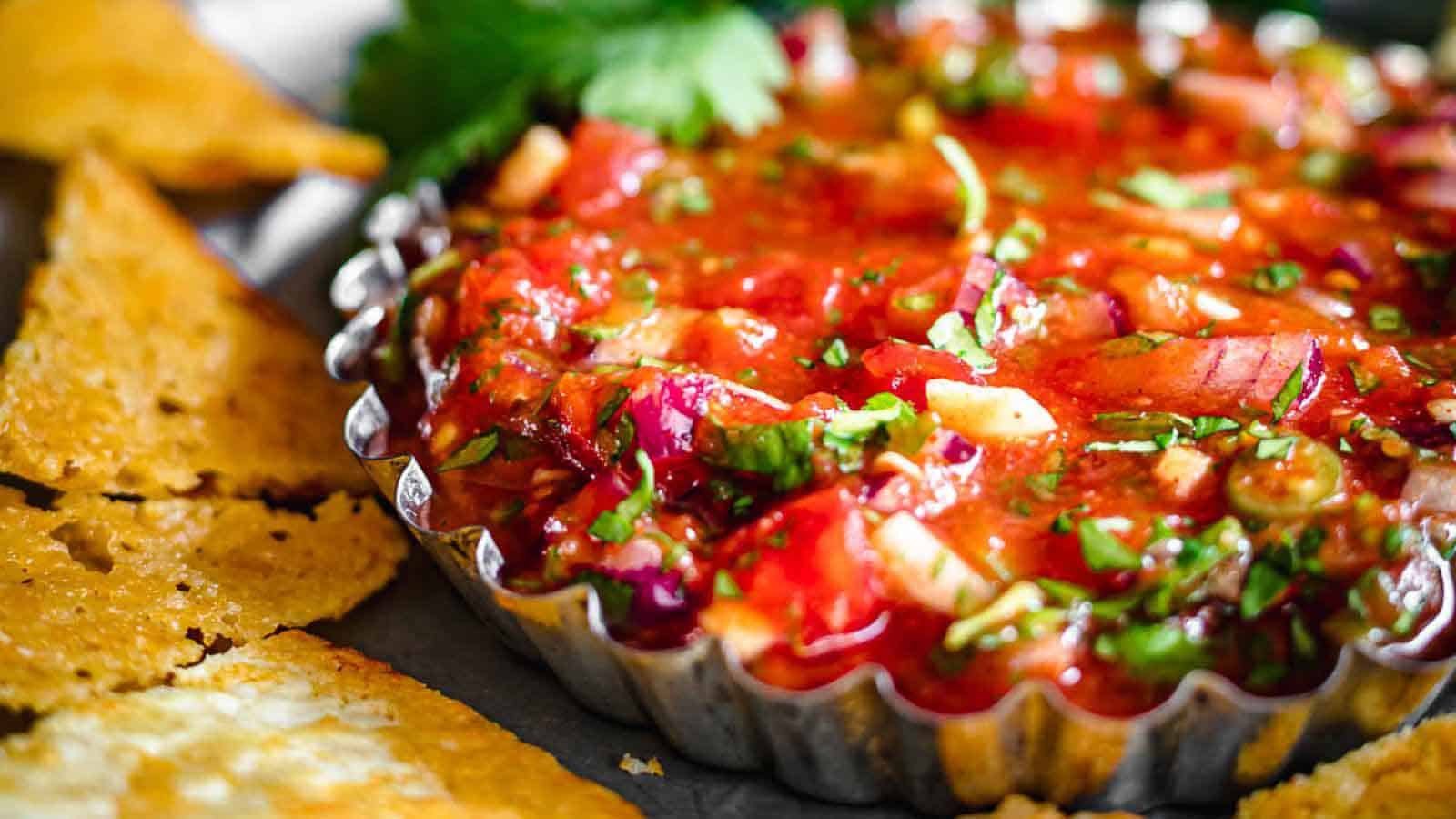 Close-up of a dish filled with fresh, vibrant salsa, featuring diced tomatoes, onions, herbs, and spices. The salsa is served in a scalloped-edge metal container, surrounded by crispy tortilla chips. A sprig of cilantro is visible in the background.