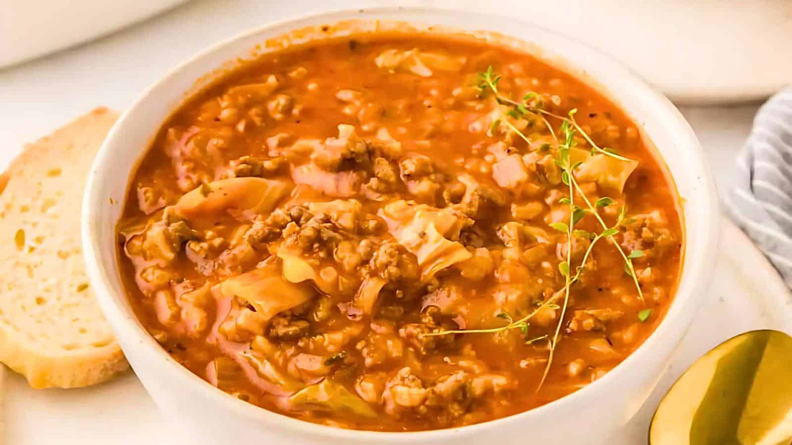 A bowl of hearty cabbage roll soup filled with ground meat, rice, and cabbage chunks, all simmered in a rich tomato-based broth. Garnished with a sprig of fresh thyme, the soup sits on a white surface next to a slice of bread and a gold-colored spoon.