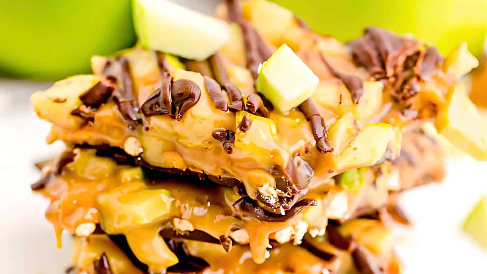 A close-up of a stack of caramel and chocolate-covered apple slices. The slices are drizzled with caramel and chocolate, with small chunks of green apple on top, showcasing a gooey, rich, and decadent treat.