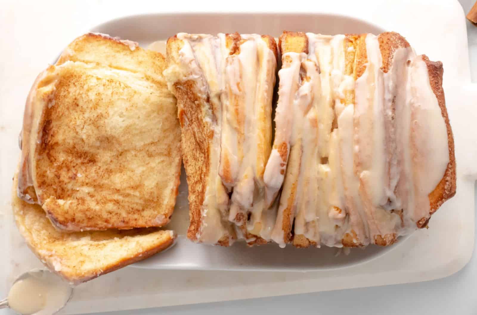 A close-up of a loaf of cinnamon pull-apart bread on a white plate. The bread is sliced and drizzled with a generous amount of white icing, with one piece slightly separated, revealing the swirled cinnamon layers inside.