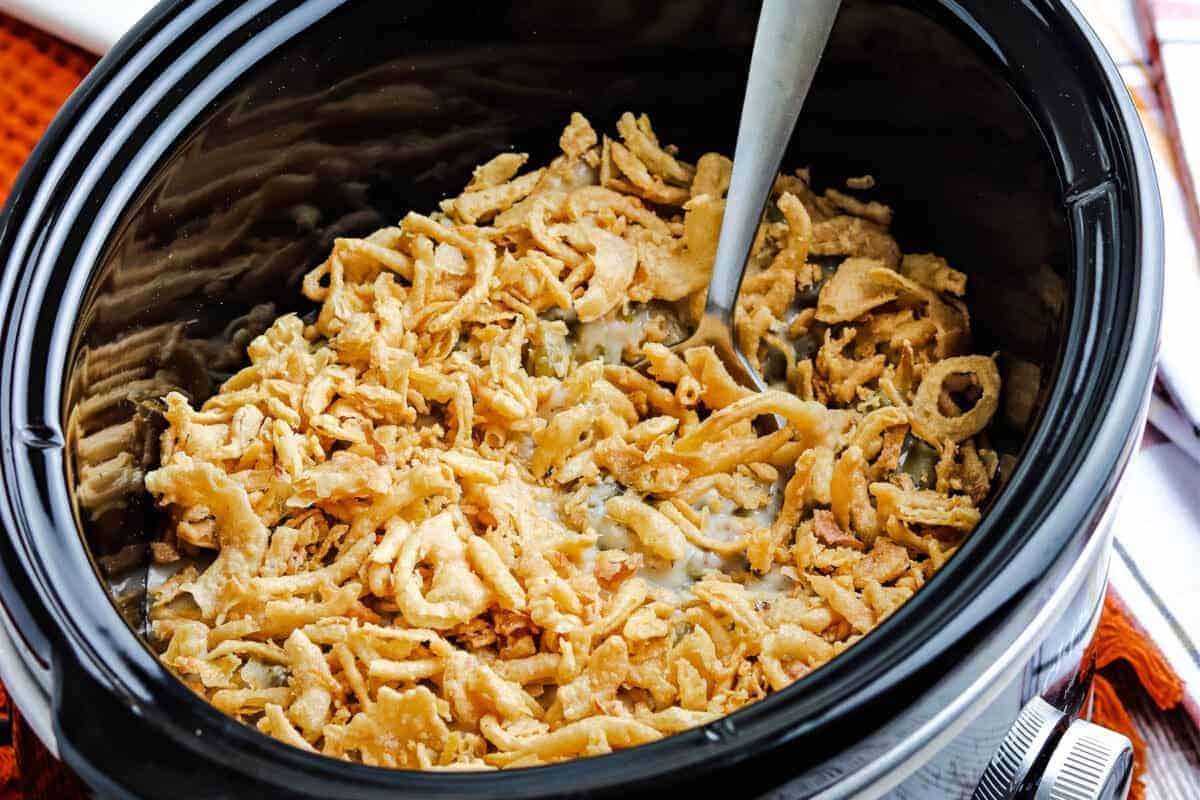 A close-up view of a slow cooker filled with a creamy casserole topped with crispy fried onions. A serving spoon is resting inside, ready to scoop out the dish. The slow cooker rests on a tabletop with part of an orange cloth visible.