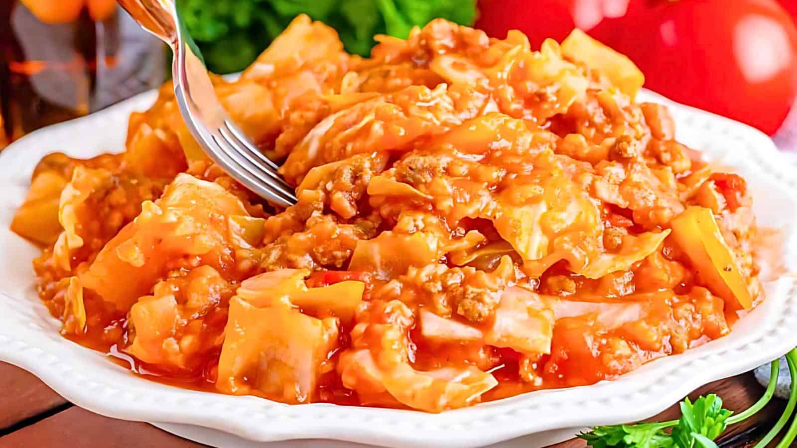 A plate of stuffed cabbage casserole featuring tender cabbage, browned ground meat, and a savory tomato sauce. The dish is garnished with fresh parsley and surrounded by ripe tomatoes and other vegetables in the background. A fork is partly holding a portion.