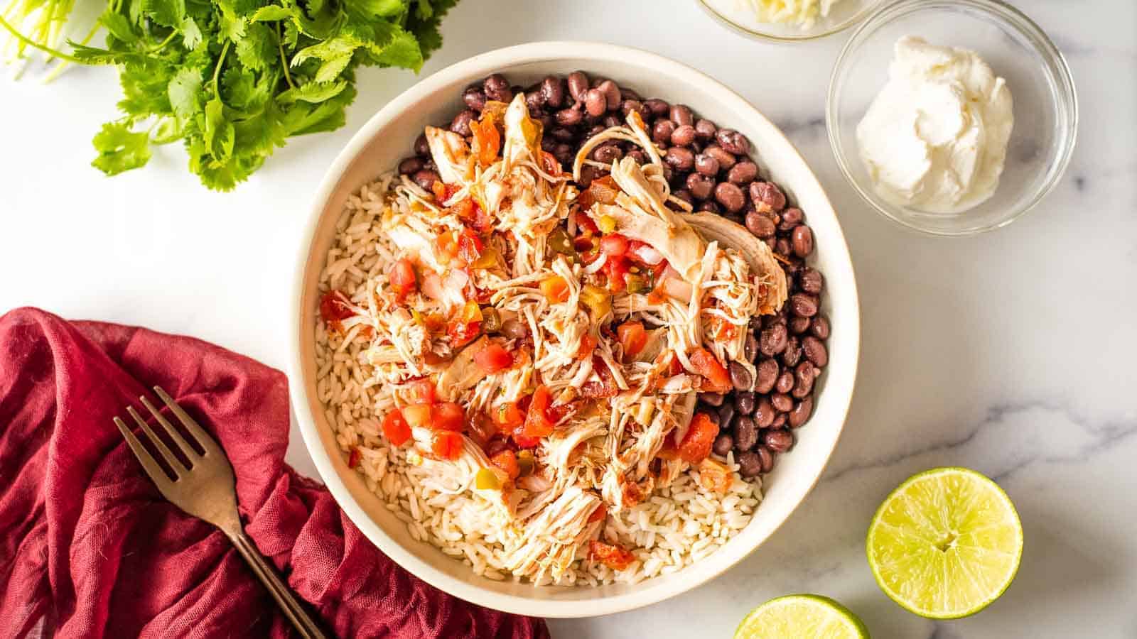 A bowl of rice topped with shredded chicken, diced tomatoes, and black beans. Surrounding the bowl are fresh cilantro, lime halves, a red cloth with a fork, and small bowls of sour cream and another sauce or dip on a white surface.
