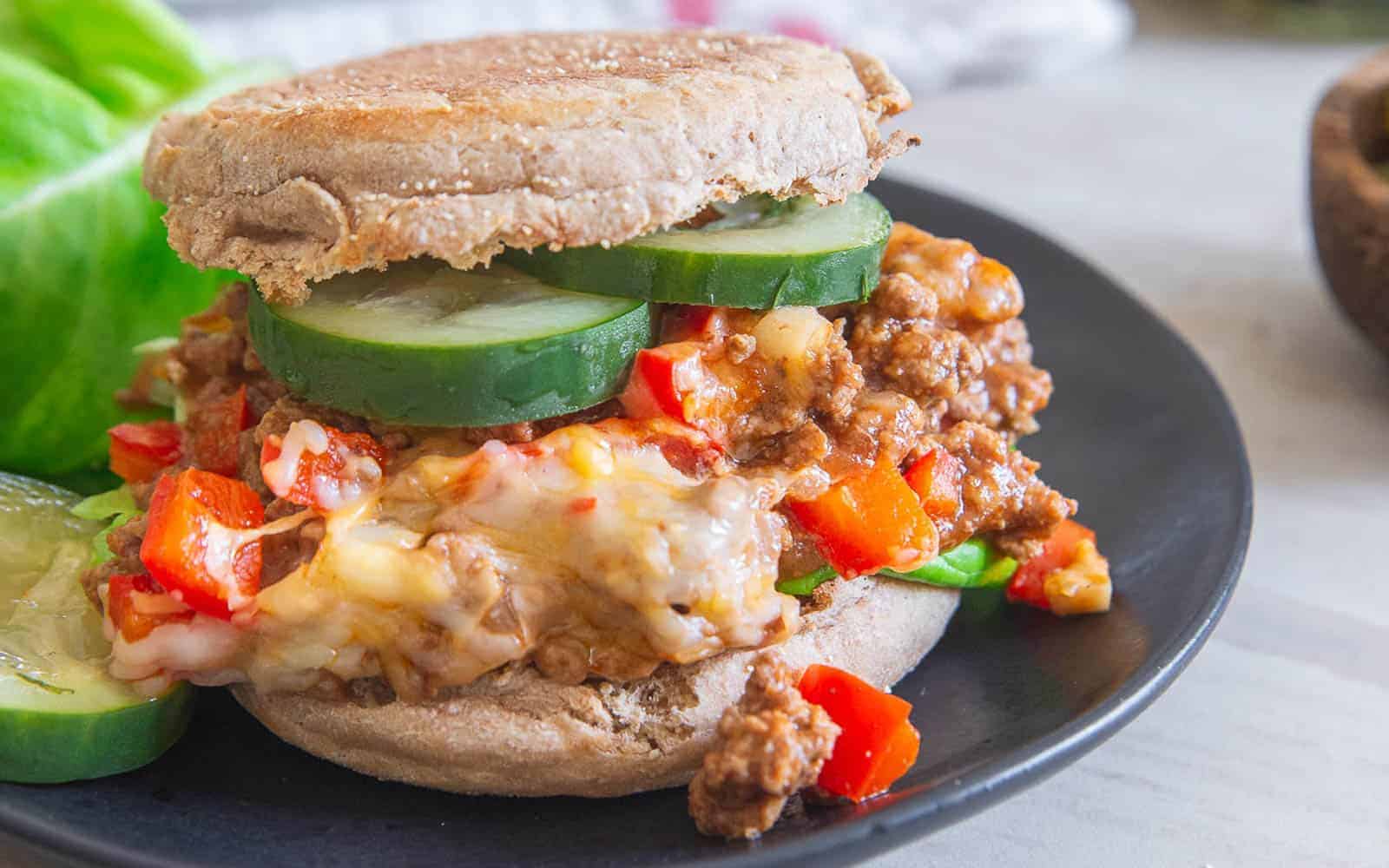 A juicy sloppy joe sandwich on a whole wheat bun sits on a black plate. The sandwich is filled with ground meat, diced red bell peppers, melted cheese, and slices of fresh cucumber. A piece of lettuce and a cucumber slice are placed next to it on the plate.