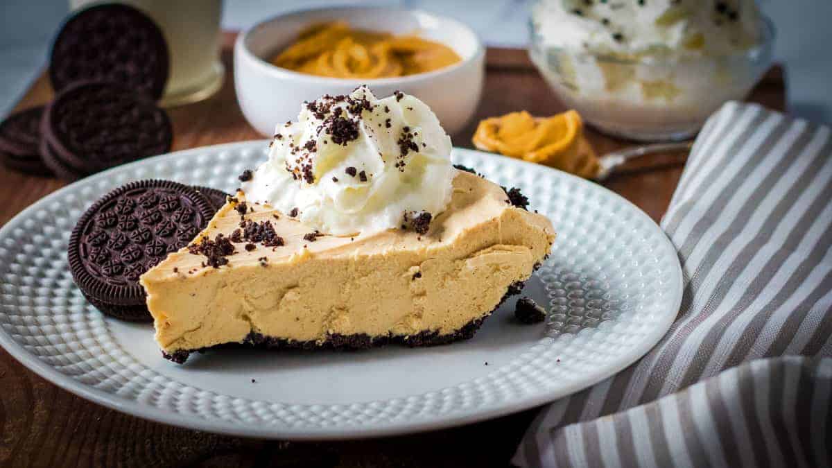 A slice of creamy peanut butter pie topped with whipped cream and chocolate cookie crumbs sits on a white plate. Next to the pie slice are two chocolate sandwich cookies. In the background are a bowl of peanut butter, more whipped cream, and a striped napkin on the side.
