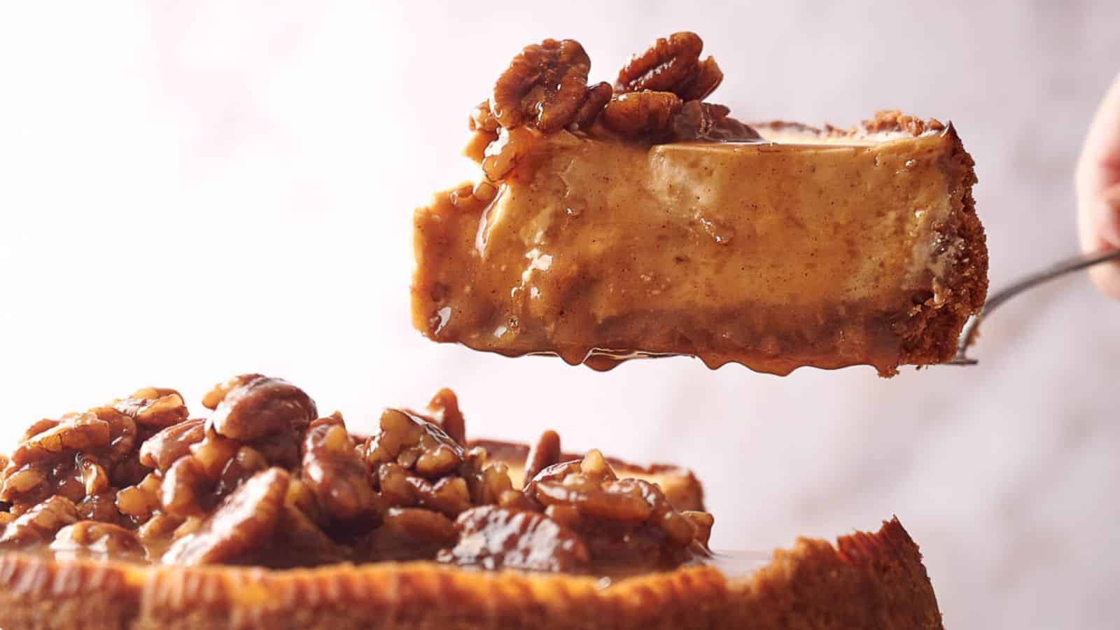 A close-up of a slice of caramel pecan cheesecake being lifted from the whole cake. The cheesecake is topped with a generous layer of glazed, chopped pecans, and the caramel sauce is dripping slightly from the slice. The background is softly blurred.