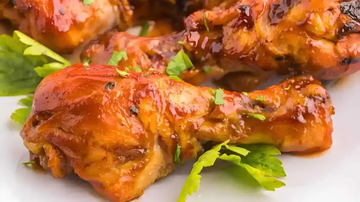 Close-up of glazed and roasted chicken drumsticks garnished with fresh parsley, placed on a white plate. The chicken looks juicy and well-seasoned, with a shiny surface indicating a flavorful sauce.