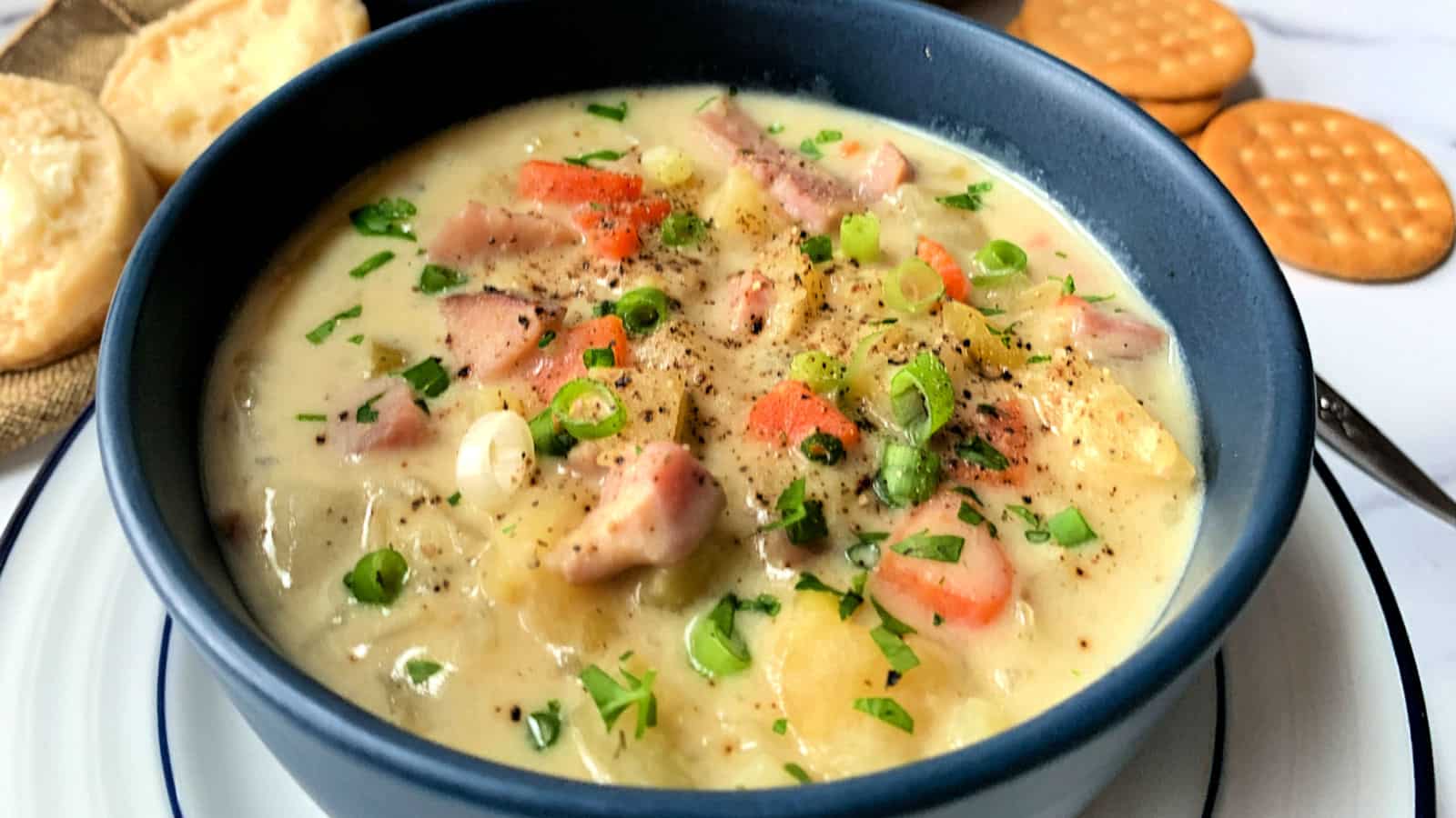 A blue bowl filled with creamy chicken and vegetable soup, garnished with chopped green onions and parsley, is placed on a white plate. Beside the bowl are round, buttery crackers on a wooden surface.
