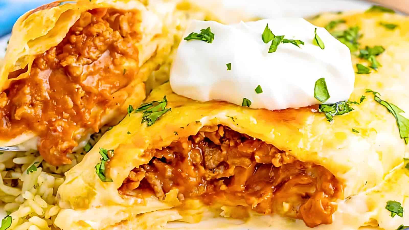 A close-up of a cheesy burrito cut in half, revealing a spicy, saucy ground meat filling. The burrito is topped with a dollop of sour cream and garnished with chopped fresh herbs, served on a bed of rice.