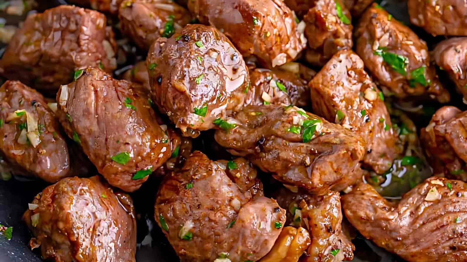 Close-up of sizzling garlic steak bites in a pan, glistening with oil and garnished with chopped parsley. The juicy, tender-looking meat chunks are coated in a flavorful marinade, showcasing a mouth-watering, golden-brown sear.