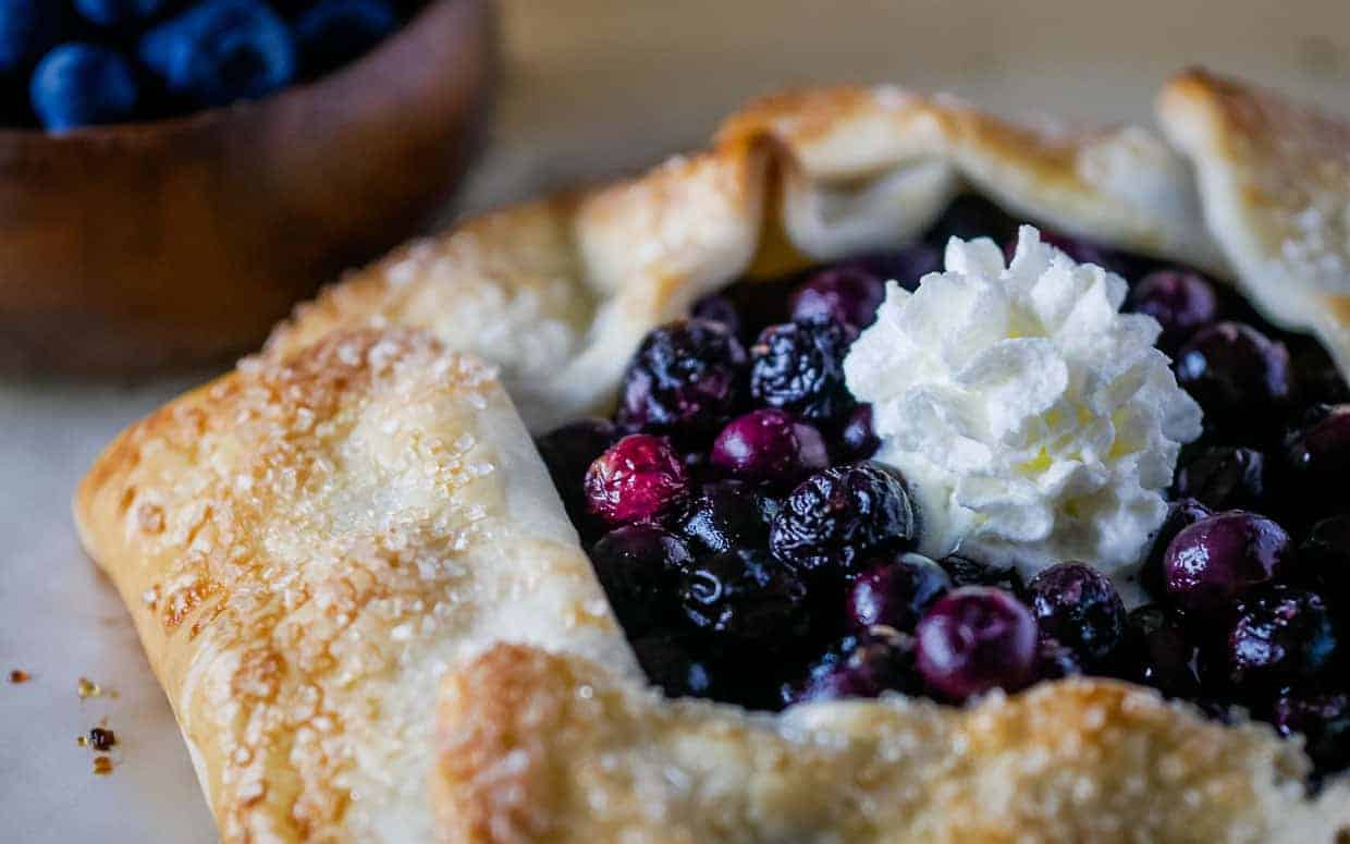 A close-up of a blueberry galette with a dollop of whipped cream in the center. The galette features a golden, flaky crust sprinkled with coarse sugar, and is filled with glistening, dark blueberries. In the background, there's a wooden bowl filled with fresh blueberries.