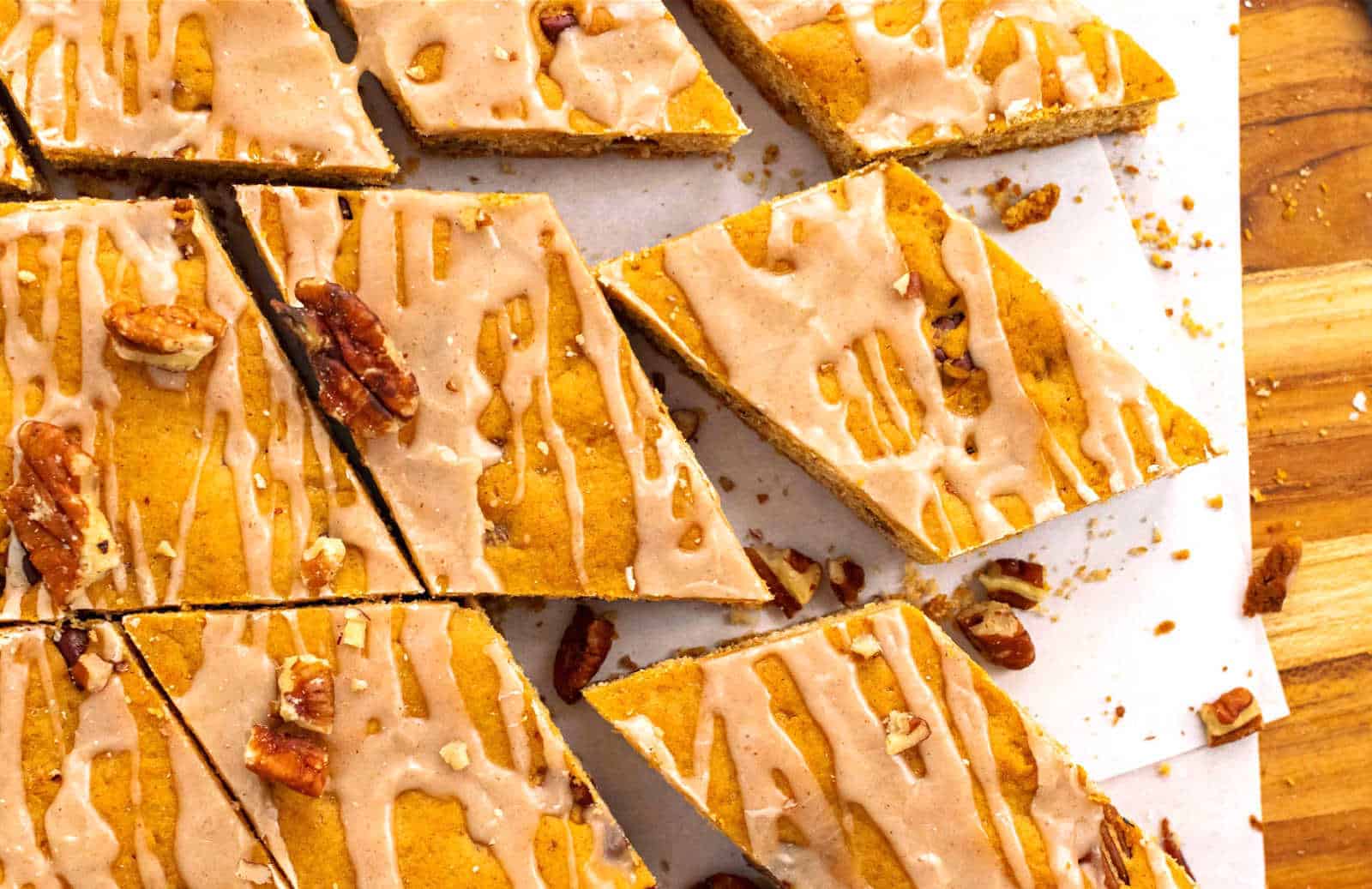 An overhead view of pumpkin bars cut into triangles, drizzled with icing and topped with pecan pieces. They are arranged on a white parchment paper over a wooden surface.