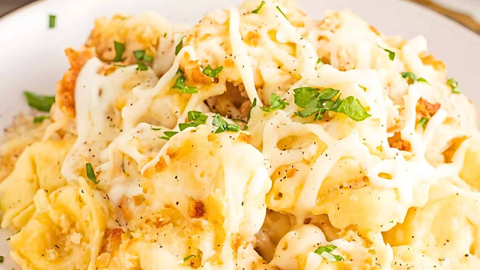 A close-up view of a plate of creamy tortellini, topped with melted cheese and garnished with finely chopped parsley. The pasta appears rich and indulgent, with a golden-brown crust suggesting it has been baked.