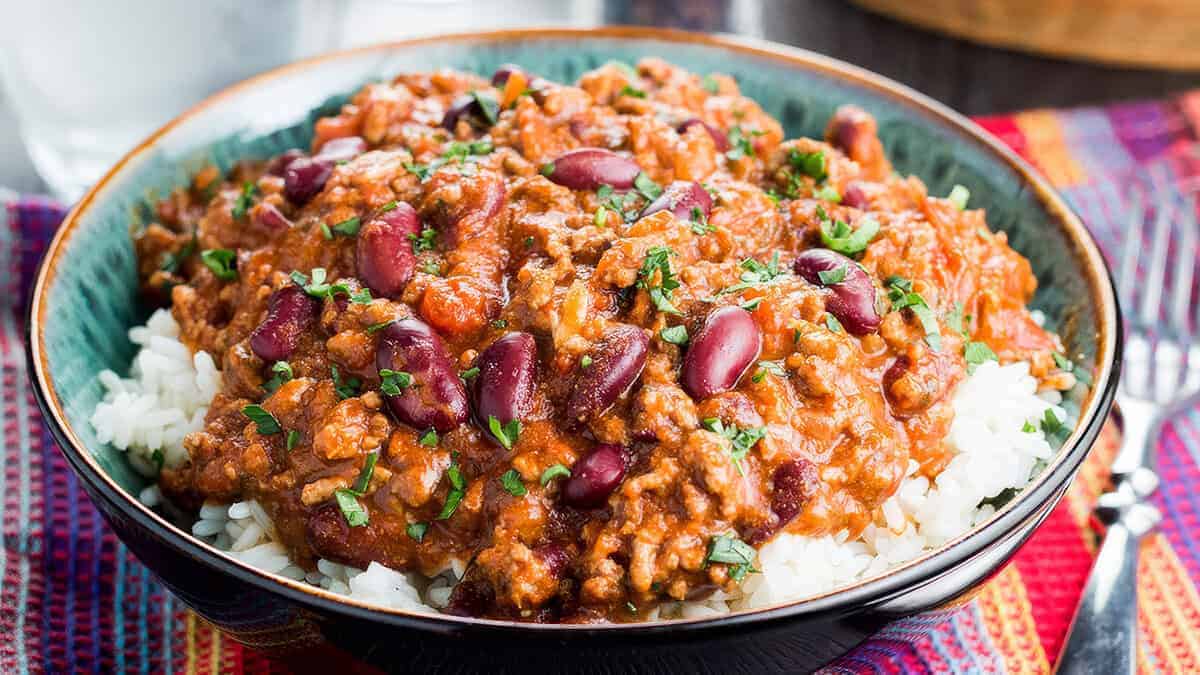 A bowl of rice topped with a generous serving of chili con carne, featuring minced meat, red kidney beans, and a rich tomato-based sauce. The dish is garnished with fresh chopped herbs and sits on a colorful woven placemat.