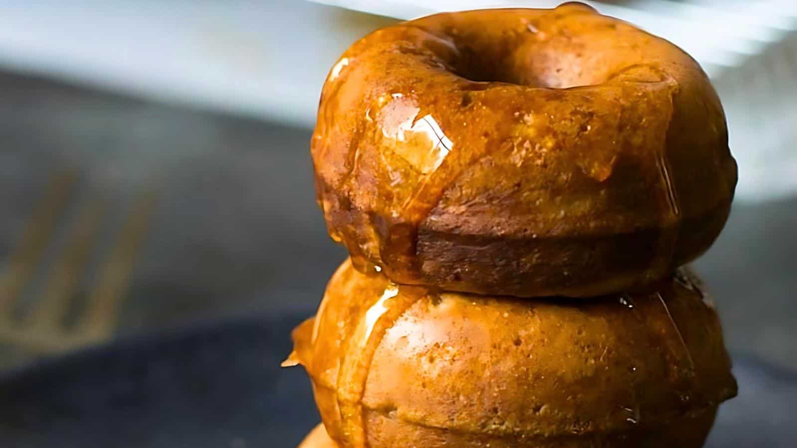 Close-up of two glazed donuts stacked on top of each other, with the shiny glaze dripping down the sides. The background is blurred, emphasizing the texture and sheen of the donuts in the foreground.
