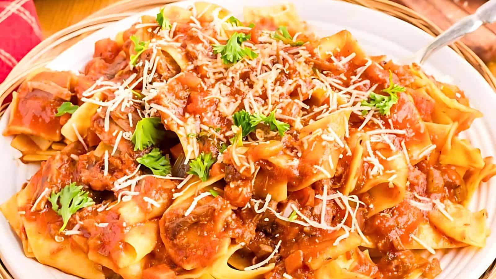 A close-up of a bowl of wide pappardelle pasta topped with a generous amount of rich meat sauce. The dish is garnished with fresh parsley and shredded Parmesan cheese, creating a vibrant and appetizing presentation.